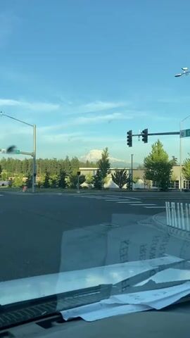 The video shows a view from inside a vehicle, looking out through the windshield. Outside, there is a street intersection with a traffic light and a street sign that reads "53 Ave E". Across the street, there is a building with a white roof and siding, surrounded by trees. In the distance, a snow-capped mountain is visible. The sky is blue with some scattered clouds. The windshield has some papers on it, including what appears to be a printout with text on it. The sun is shining, and the lighting suggests it is either early morning or late afternoon. There are no people visible in the video. The scene appears to be in an urban or suburban area with natural scenery in the background.