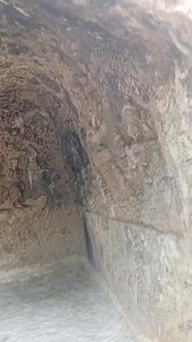 The video begins showing the interior of a cave or rock shelter with rough, carved stone walls. The camera then pans to a man standing outside the shelter, looking at the carvings. The scene transitions to an outdoor shot of a path leading through a wooded area with trees. There is a small structure with a tiled roof on the left and a sign in the background. The man from the earlier shot is partially visible on the right side of the frame.