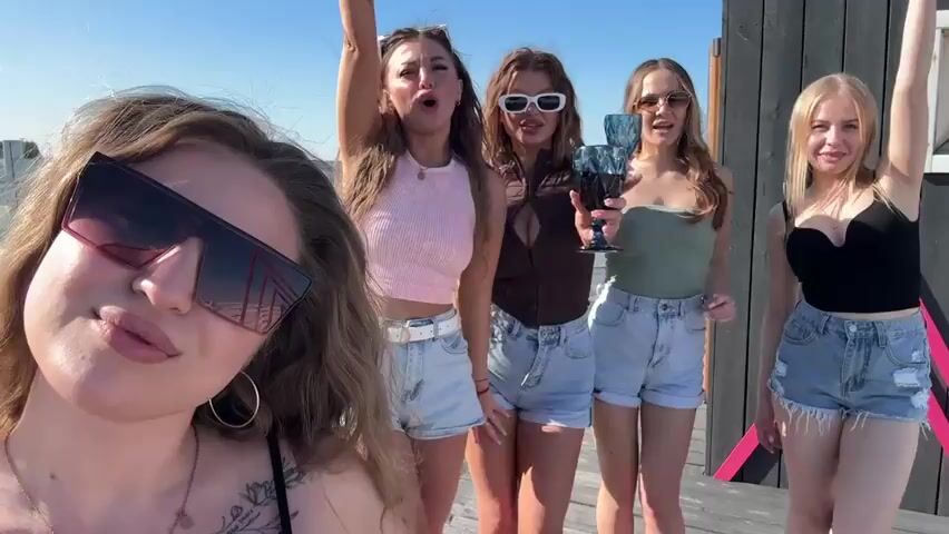 The video depicts five young women in a sunny outdoor setting. They are standing close together, with one woman taking a selfie-style video, partially obscuring the left side of the frame. The other four women are posed behind her. Each of the women is wearing casual summer attire, including shorts and tank tops. Most of them are holding a blue stemmed glass, and they appear to be toasting or celebrating. The background includes a wooden structure with pink accents, suggesting a beachside or lakeside setting. The women are smiling, appearing happy and relaxed.