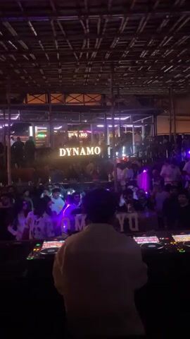 A DJ stands behind a console, facing a crowd gathered in a dimly lit venue. The ceiling is adorned with a lattice of wooden beams, and a neon sign spelling "DYNAMO" illuminates the space. People are seen dancing and socializing, illuminated by colorful stage lights.