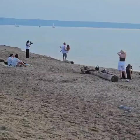 The video shows a beach scene with several people. In the foreground, the sandy beach is visible. Further back, on the shoreline, a man is holding a woman in his arms, and they appear to be posing for a photo. A woman with a camera is standing to their left, apparently taking their picture. To the left of the couple, two people are sitting on the sand, facing away from the camera. To the right of the couple, a woman in a black top and white shorts is standing and looking out at the water. A large log lies on the sand between the couple and the standing woman. In the background, a body of water is visible, with several sailboats in the distance. The sky is overcast and a hazy, distant shoreline can be seen.
