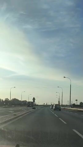 The video shows a view from the driver's seat of a car on a highway under a partially cloudy sky. The road has multiple lanes and streetlights on either side. There are other cars and a truck visible on the road. The sun is low in the sky, casting a warm light. A speed limit sign of 50 is visible on the right side of the road. The video pans slightly to the right and then returns to a forward-facing view.