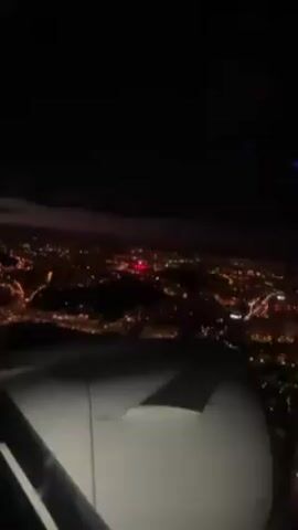 The video shows an airplane approaching a city at night. The city is illuminated by many lights, appearing as a dense network of orange and white dots. A prominent red light is also visible. The airplane's wing and engine are partially visible in the foreground. The sky is dark, with some cloud cover faintly illuminated by the city lights.
