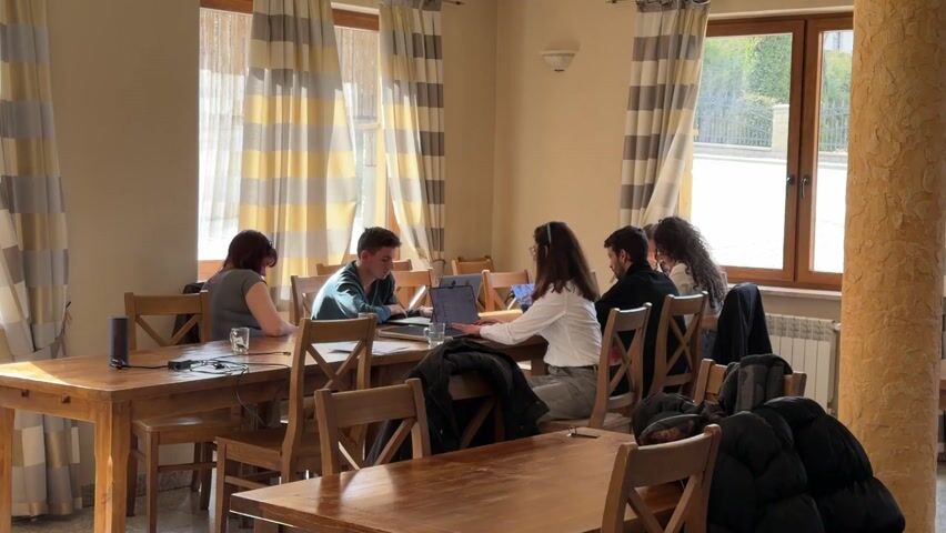 Several people are gathered in a bright room, seated at wooden tables. Three windows adorned with striped curtains allow sunlight to illuminate the space. Most of the individuals are focused on laptops or papers spread out before them, seemingly engaged in work or study. Coats are draped over the backs of the chairs. The room has a warm, casual atmosphere, suggesting it could be a common area in a lodge or retreat.