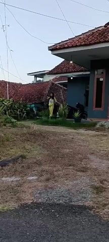 A person in yellow pajamas is standing in front of a house. The person then walks away from the camera and disappears behind a motorcycle. The scene then changes to a wider shot of a village in a hilly, forested area. Several houses with red tile roofs are visible, along with lush green vegetation. The sky is overcast.