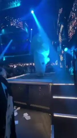 The video shows a nightclub with a stage where a person in an inflatable white mushroom costume is standing. The stage is lit with blue spotlights. The club is crowded with people. The camera is positioned near the stage, and someone wearing a patterned black-and-white shirt is partially visible in the foreground. There are some white tissues on the floor.