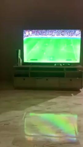 A low-angle shot captures a living room. A large screen displays a soccer video game. Underneath the TV is a light-colored wooden TV stand. A PlayStation 5 sits to the left. There are game controllers on the stand. A multicolored light reflects on the marble floor. Music plays.