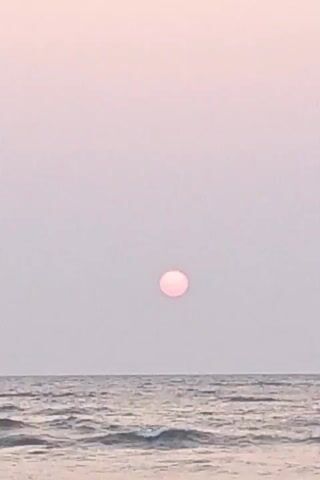The video presents a wide shot of a calm sea under a clear, soft pink sky. The sun, appearing as a small, light pink orb, is centered in the upper part of the frame. Gentle waves ripple across the sea, with the horizon line positioned in the lower third of the view. No people or objects are discernible beyond the natural elements. The setting suggests a serene moment at sunrise or sunset.