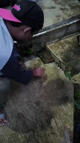 A person in a black baseball cap with a pink brim is crouching on a concrete surface. The person is wearing a gray long-sleeved shirt with a blue underlayer, khaki pants, and gray sandals. They are holding a clear glass in their right hand and appear to be sniffing the contents before taking a sip. They then turn towards the camera and smile broadly, holding up the glass. The concrete surface has a grooved texture and there is some greenery growing nearby. A box of cigarettes and a lit cigarette are visible on the concrete surface. The scene is dimly lit.