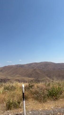 The video pans across a dry, mountainous landscape under a clear blue sky with scattered clouds. The terrain is characterized by rolling hills covered in dry, golden vegetation and sparse green shrubs. In the foreground, there is a rough, gravelly surface. A white and red road marker is visible on the right side of the frame in the first few seconds. In the distance, more rounded, barren mountains stretch across the horizon. Towards the middle of the video, some rock formations and shadows suggest ravines or depressions in the landscape. In the third and fourth seconds, a dark, angular structure, possibly an old ruin or fence post, appears on the left side of the frame.