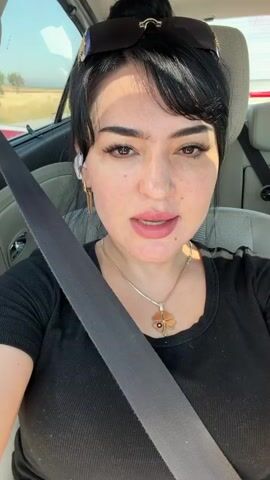 A woman with dark hair pulled into a bun, wearing sunglasses on top of her head, a black t-shirt, and a necklace with a clover pendant is sitting in the driver's seat of a car. She is wearing a seatbelt. She is looking at the camera and speaking. The interior of the car is visible, including the passenger seat and the car's dashboard. Outside the car window, another vehicle and a road are visible. The sun is shining.