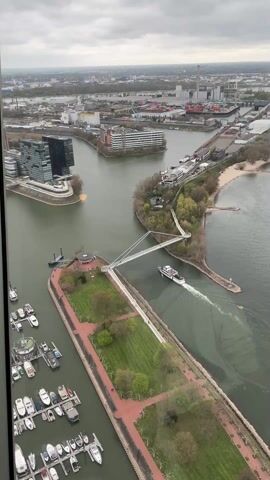 The video shows an aerial view of a river with a cruise boat passing under a modern suspension bridge. The boat, predominantly white with some pink trim, is moving from right to left, creating a wake in the green water. The bridge is a minimalist design with a gray support structure and a white walkway. People can be seen walking on the bridge. 

The scene includes land on both sides of the river with green grass, trees, and red brick walkways. On the left, there is a marina with numerous docked boats. In the background, the cityscape includes modern buildings and industrial areas. The sky is overcast, suggesting a cloudy day. The camera zooms in slightly on the boat as it passes under the bridge.