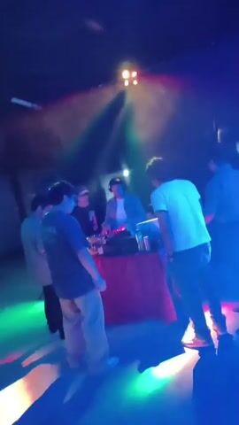The video shows a DJ playing music for a small group of people in a dimly lit club. The DJ is positioned behind a DJ booth equipped with audio equipment, and he is wearing headphones. The group of people are standing in front of the booth, seemingly enjoying the music. The club is illuminated by colorful spotlights, creating a vibrant atmosphere. One of the individuals in the group is wearing a shirt that says "REVIVAL".