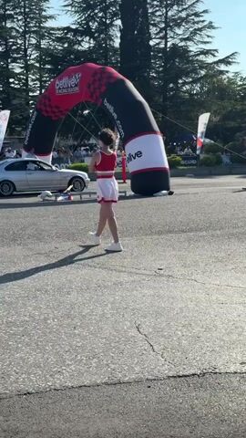 The video shows a street race event. A female presenter in a red and white outfit stands near an inflatable archway with the logo "betlive RACING SEASON". A silver car is positioned behind a small platform. In the background, spectators are gathered, and trees and buildings are visible. The overall environment appears to be an outdoor urban setting.