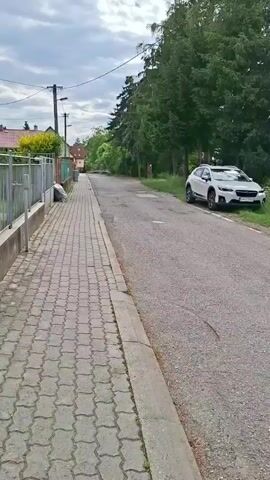 A white Subaru XV is parked on the right side of a paved road, near trees and a grassy embankment. A paved sidewalk runs along the left side of the road, with a metal fence bordering it. In the distance, a house with a red roof is visible, along with more trees and a utility pole. The sky is cloudy.