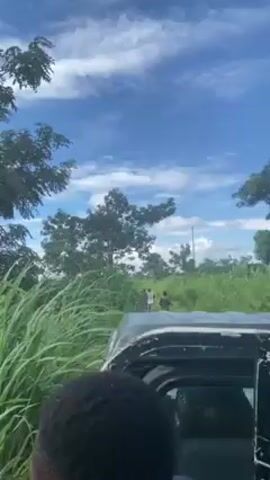 The video shows the perspective from a vehicle driving down a dirt path. The path is surrounded by tall grass and trees. Two people are walking ahead of the vehicle on the path. The sky is blue with white clouds.