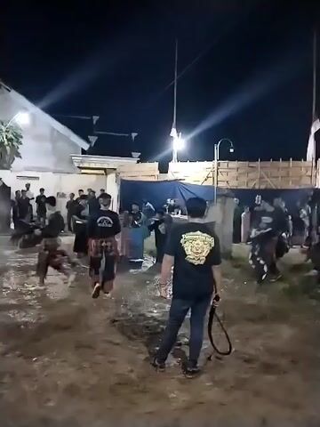 The video captures a nighttime cultural performance taking place outdoors on a dirt and muddy ground. Numerous individuals, predominantly men, are present, with several engaged in a dynamic, martial arts-like dance or ritual. Two performers in the foreground are actively moving, one striking the other who then falls to the ground. In the immediate foreground, a man, viewed from behind, holds a whip and observes the scene. He wears a black t-shirt with a prominent golden crest-like design on the back, featuring what appears to be a mythical creature's head and text that includes "JATHILAN" and "JENGGRIK".

The setting includes simple, lit buildings in the background and temporary structures made of bamboo and a blue tarp. Artificial lights illuminate the area, suggesting an organized event. Other people are standing around, watching the performance. The muddy ground indicates recent wet weather or damp conditions. The overall atmosphere appears focused and energetic.