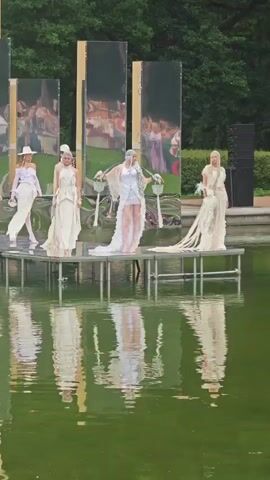 This video captures a fashion show taking place outdoors in Pushkin, Russia. A runway has been set up over a body of water, creating a reflective surface for the models and the backdrop. The models are walking one by one, showcasing a collection of white and light-colored garments, many with flowing fabrics and elaborate designs. Some models are wearing headpieces, adding to the ethereal and perhaps bridal aesthetic of the collection.

Behind the runway, large, mirrored panels are erected vertically, reflecting the surrounding environment and the models themselves. In the background, a grand, pale yellow building with arched windows is visible, suggesting a historical or palatial setting. Trees and foliage form a lush green backdrop, indicating it is likely daytime and the weather is fair. The overall atmosphere appears sophisticated and artistic, with the water and mirrors adding a unique visual dimension to the fashion presentation. The reflections on the water are particularly striking, mirroring the models and the architecture. No visible text is present in the video.
