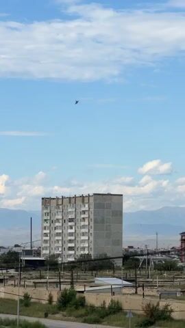 A fighter jet flies across a blue sky with scattered white clouds. Below the jet, a tall, concrete apartment building stands in a semi-urban area. In the background, mountains are visible under the horizon. The camera appears to be stationary, looking upwards at the sky.