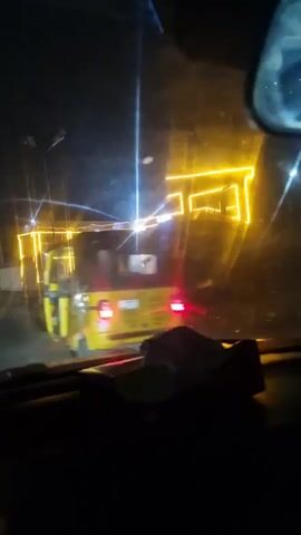 The video shows a drive at night, presumably through a town. The camera is inside a car, pointed through the windshield. Bright yellow neon lights outlining a building are seen at the start, followed by the car driving past it.  The street is wet. The camera then shows the car's dashboard, including the speedometer. The car's speed fluctuates, with the needle swinging around the 20 mph mark. Warning lights are illuminated on the dashboard. Other cars pass by in the opposite direction.
