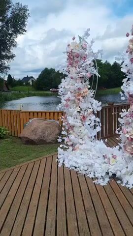A video displays a large, ornate floral archway, decorated with white and pink flowers and greenery. The archway stands on a wooden deck, with a body of water and trees in the background. The sky is cloudy. A large rock is visible to the left of the archway.