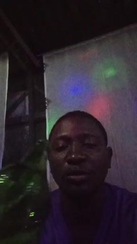 A man with dark skin and a purple shirt is drinking from a green glass beer bottle. He is indoors, and there are colored lights shining on a white curtain behind him. The bottle is labeled "TYCKA". There is music playing in the background.