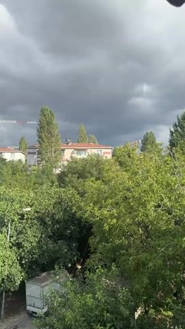 The video shows a panoramic view of a residential area with buildings and lush green trees. The sky is overcast with dark, heavy clouds, suggesting an impending storm or rainy weather. The camera pans across the scene, revealing apartment buildings with balconies and windows. In the foreground, the foliage is dense, with the leaves of various trees casting shadows. A white delivery truck with a license plate is partially visible on the left side of the frame. In the background, construction cranes can be seen, indicating ongoing development in the area. The overall atmosphere is somber due to the cloudy sky, but the greenery provides a sense of life and nature.
