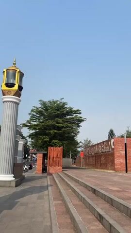 The video pans to the right, capturing an outdoor public space under a clear blue sky. In the foreground, a paved walkway leads to a set of concrete steps. To the left, a large, fluted column with a decorative lantern-like fixture stands. Beyond the column, a street with parked motorcycles is visible.

The middle ground features a large tree with dense green foliage, situated behind a small, modern structure constructed with brick. Adjacent to this structure is a long, low wall made of brick, with lettering etched into its surface. The wall leads to a modern building with an open entrance, also constructed primarily of brick. Several black lampposts with rectangular white lights are positioned around this area.

Further to the right, the space opens up to reveal tiered seating made of brick, resembling an amphitheater. More lampposts are scattered throughout this section. The background consists of more trees and a distant building. The overall impression is of a well-maintained public plaza or park.