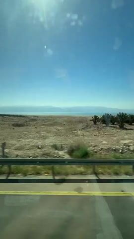 The video shows a view from inside a moving vehicle on a sunny day. The road is divided by a yellow line, and a metal guardrail is visible along the edge. Beyond the road, there is a flat, arid landscape with sparse vegetation. In the distance, a body of water is visible, likely a lake or sea, with hills or landmasses on the far shore. The sky is clear and blue. There are several date palm trees planted in the landscape. The sun is bright and causes glare in the camera.
