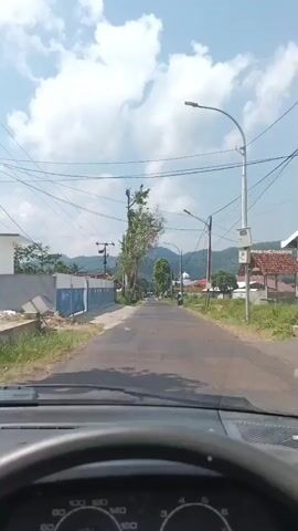 The video shows a view from inside a car driving down a paved road. The road is lined with trees and houses on either side. The sky is blue with white clouds. There is a lamppost and power lines visible. In the distance, there are hills. The dashboard of the car is visible at the bottom of the frame.