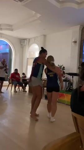 Two women in athletic attire are dancing barefoot in a room with light-colored wooden floors. The woman on the left has dark hair pulled back and is wearing a blue and teal athletic top with gray shorts. She is smiling and appears to be in motion. The woman on the right has blonde hair and is wearing a similar athletic top and black shorts. She is also smiling and appears to be dancing. In the background, several other people are visible, some seated and others standing. The room is well-lit with overhead lighting and appears to be a living or common area, with furniture and what looks like a stage or elevated platform with equipment.
