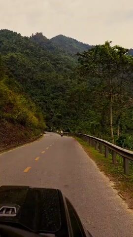 The video shows a view from the front of a car driving on a winding road through a mountainous, forested area. The road is paved with yellow lane markings and a metal guardrail along the right side. Lush green vegetation covers the steep slopes of the mountains on both sides of the road. In the distance, a few other vehicles, including a red car and a moped with two riders, can be seen traveling in the same direction. The sky is overcast. A road sign and kilometer marker are visible along the side of the road. The hood of the car, slightly dirty, is visible at the bottom of the frame.