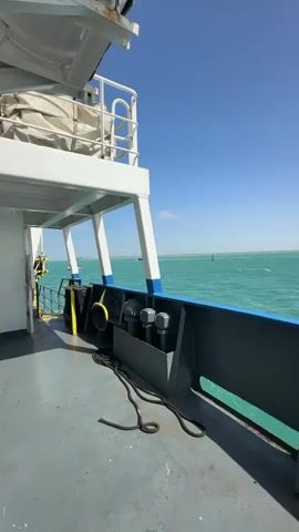 A tugboat passes a larger vessel on a clear, sunny day. The video is filmed from the deck of the larger vessel, looking out over calm, turquoise-colored water. The tugboat, primarily white and grey with a tall mast, moves from right to left across the frame, creating a visible wake. In the foreground, parts of the deck are visible, including a yellow-topped black bollard, coiled ropes, and a grey panel with "2.0M" written on it. Overhead, a white structure with railings and a covered object (possibly a life raft) is visible. The sky is a bright, cloudless blue. A distant coastline or landmass can be seen on the horizon. The sound of a low hum or engine noise is present.