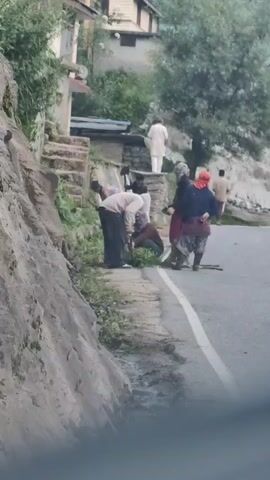 Several people are gathered on the side of a road next to a rocky incline. Two individuals in the foreground are bent over, appearing to clear vegetation from the roadside. One person is wearing a light-colored shirt and dark pants, and the other is wearing a darker shirt and appears to be assisting. Behind them, two women are standing, one in a red scarf and dark clothing, and another in a dark headscarf and a patterned red garment. To the right, a person in a light-colored shirt and pants is walking away from the group. Further in the background, a person in a white garment is standing on a raised platform. The environment includes lush green trees and vegetation on a slope behind the people, and buildings are visible on the hillside. The road has a white dividing line.