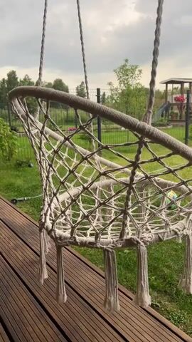 The video shows a close-up of a macrame swing chair hanging from ropes. The chair is made of thick, woven rope and has a net-like structure. It is hanging over a wooden deck, with lush green grass and trees visible in the background. A fence separates the yard from a wooded area. In the distance, a playground structure with a slide and a small red roof can be seen. The sky is overcast, suggesting a cloudy day. The overall mood is peaceful and relaxing, evoking a sense of leisure and outdoor enjoyment. The macrame chair itself is a focal point, highlighting its intricate craftsmanship and comfortable design, suggesting it's a perfect spot for relaxation. The presence of the playground implies a family-friendly setting, possibly a backyard or garden area. The time of day appears to be daytime, given the natural light, but the cloudy sky obscures any specific indication of morning, afternoon, or evening. There is no visible text in the media.