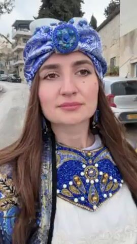 A woman with long brown hair is walking down a street in what appears to be a Middle Eastern town. She is wearing an elaborate blue and gold beaded head covering and matching beaded collar over a white blouse. She is wearing a matching jacket. The street is narrow with cars parked on both sides. The buildings are old and made of stone. The sky is overcast. The woman is looking at the camera and smiling slightly. The video appears to be a selfie.
