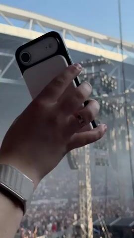 The video shows a large outdoor concert with a massive crowd of people in an open-air stadium. In the foreground, a person's hand holds a smartphone attached to a white battery pack, suggesting they are recording or taking pictures of the event. The camera is pointed towards the stage area where a complex lighting rig and speaker system are prominently displayed. Smoke or fog effects are visible around the stage and emanating upwards. The stadium seating is packed with spectators, and the overall atmosphere appears to be energetic and lively. The lighting is bright, indicating it is daytime or the stadium lights are very powerful.