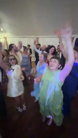 The video shows a lively wedding reception inside a tent. Several people are dancing and enjoying themselves, with some raising their hands in the air. In the background, a live band is performing on a stage adorned with flowers, featuring a singer and instrumentalists. The lighting is a mix of warm string lights and vibrant colored stage lights, creating a festive atmosphere. People are dressed in formal and semi-formal attire, including dresses and suits. The overall scene depicts a joyful celebration.