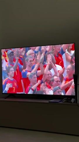 A flat screen television displays a broadcast of a soccer match, showing a crowd of fans wearing red and yellow. The fans are clapping and cheering, with many wearing red jerseys with yellow accents. The atmosphere appears festive and energetic. On the shelf below the television, a black video game console and a controller are visible. The room is dimly lit, suggesting it is evening or the blinds are drawn. The overall scene depicts a group of people enthusiastically watching a sporting event.