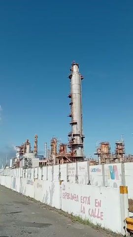 The video presents a wide shot of an industrial complex, likely an oil refinery, under a clear blue sky with scattered clouds. The foreground features a concrete wall, decorated with graffiti slogans and corporate logos, running along a paved road. In the background, a sprawling network of metallic structures, including tall distillation columns, pipes, and various processing units, dominates the landscape. Some units are releasing plumes of smoke, suggesting ongoing operations. The overall impression is of a large-scale industrial facility engaged in refining or processing raw materials. The camera appears to be in a moving vehicle, travelling alongside the facility.