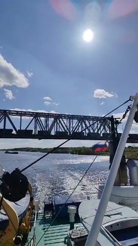 A water vessel moving towards a large steel bridge with a train on top. The scene is viewed from the vessel, with a Russian flag visible on the vessel. The water sparkles in the bright sunlight, and there are clouds in the clear blue sky. The camera is stable and the angle is low to mid-level.