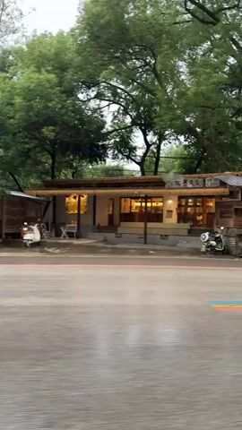 The camera, likely from a moving vehicle, captures a rainy street scene. A restaurant with a rustic design sits by the roadside, with a man standing nearby. As the camera moves, a brown car passes by. Then, a person on a scooter, using a light blue umbrella, drives away on the wet road. The surrounding area is green with trees and foliage. A rock formation can be seen in the distance above the trees.