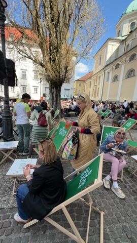 The video shows an outdoor gathering on a cobblestone square.  Many people are seated in green folding chairs, some eating from plates on small tables. Some are walking around the area.  A woman in a gold jacket walks through the chairs, carrying a colorful bag and a drink. A man in a green t-shirt stands near what looks like a small stage or vendor booth. The square is surrounded by buildings with arched doorways.  A large building with a green dome is visible in the background. The sky is blue with some clouds.