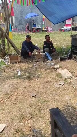 Two men are crouching on the ground outdoors, facing each other. The man on the left is wearing a dark blue long-sleeved shirt, dark pants, and a cap. He is reaching into a cardboard box in front of him. The man on the right is wearing a dark long-sleeved shirt with patches on the sleeve and dark pants. He appears to be smoking. Between them are a few more cardboard boxes, a blue cylindrical object, and some loose debris. In the background, there is a building, a blue umbrella, and various decorations including flags. The ground is covered with dry grass and dirt.