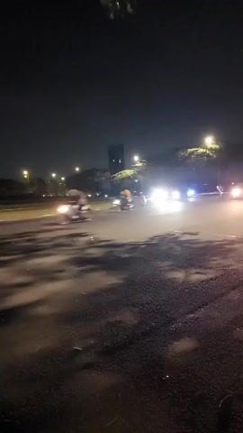 A car is seen driving at night on a road. The car appears to be a silver hatchback. There are streetlights visible in the background. The road appears to be wet, possibly from rain. There are trees on the side of the road. The video is shaky and appears to be filmed from another moving vehicle.