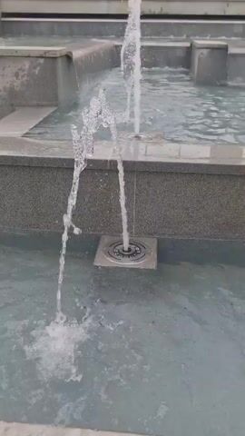 The video shows a close-up view of water jets from a fountain. The jets are spraying water upwards and then falling back into a pool. The pool appears to be made of concrete and has a textured finish. There are also steps visible in the background, which are also made of concrete. The water is clear and transparent, and the jets are creating ripples and splashes on the surface of the water. The overall scene is calm and serene, with the sound of the water providing a soothing ambiance.