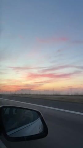 The video shows a car's side mirror reflecting the interior of the car, including a person's head and hand holding a phone. The car appears to be on a highway, as indicated by the road and the passing scenery. The sky is visible in the background, with a mix of blue and pink hues, suggesting it's either sunrise or sunset. There are some clouds in the sky. The video also shows the road ahead and the car passing by other vehicles, one of which is a dark SUV in front of the camera. The overall mood is peaceful and serene, likely capturing a moment of travel during a beautiful time of day.