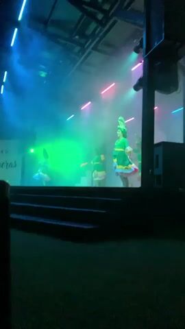 A video taken from a low angle shows a stage with performers. There are at least four people on stage wearing green dresses and elaborate headpieces. The stage is lit with green and pink lights, and there is smoke or fog filling the air. The performers appear to be dancing or acting. The stage has a raised platform in the background. The overall atmosphere is colorful and theatrical.