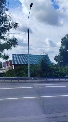 The video shows a roadside view, with a road in the foreground and a blue glass building on the right. There are trees and a house on the left. A red SUV drives by. Then a black car drifts around a curve, kicking up smoke. The sky is partly cloudy.