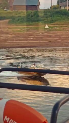 The video captures a scene on a body of water, likely a river, near Vorogovo, Russia. The foreground is dominated by the dark railing of what appears to be a larger boat or ferry, with a partially visible orange life preserver. The water is rippled, reflecting the warm, golden light of either sunrise or sunset.

In the mid-ground, a smaller, golden-colored motorboat is traversing the water, leaving a white wake behind it. A person, wearing a light-colored shirt, is seated in the driver's position of this motorboat, steering it. The boat has an outboard motor.

The background shows a gently sloping riverbank with dry, sandy soil. Further back, there is greenery, and in the distance, structures resembling rural buildings can be seen amongst the foliage. A small white triangular marker is visible on the riverbank. The overall impression is one of a serene, possibly late afternoon or early morning, excursion on a calm waterway in a rural Russian setting. No visible text is present in the video.
